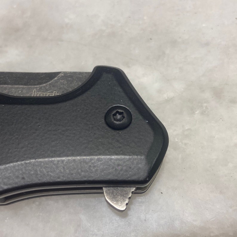 May include: Close-up of a black pocket knife with a textured handle and a silver blade. A small screw with a star-shaped head is visible on the handle.