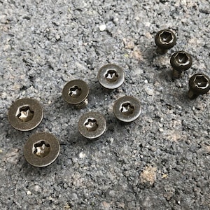 Puede incluir: Primer plano de diez tornillos de color dorado con cabeza en forma de estrella. Los tornillos están dispuestos en dos filas sobre una superficie rugosa de color gris.