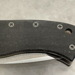May include: Close-up of a black knife handle with a textured surface and four visible screws. The handle is curved and has a slight indentation on the side.