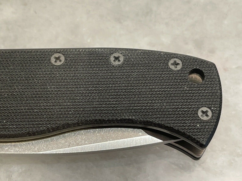 May include: Close-up of a black knife handle with a textured surface and four visible screws. The handle is curved and has a slight indentation on the side.