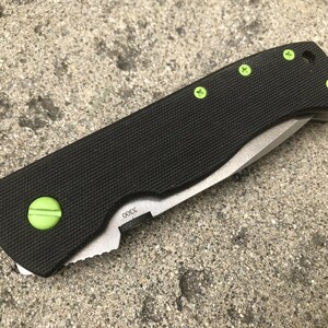 May include: A black folding knife with a green handle and green screws. The blade is partially open and the knife is sitting on a gray concrete surface.