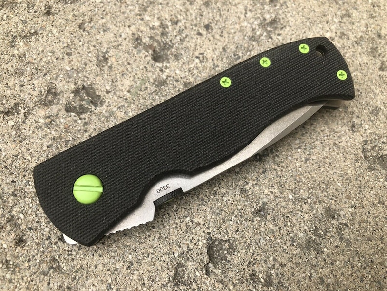 May include: A black folding knife with a green handle and green screws. The blade is partially open and the knife is sitting on a gray concrete surface.