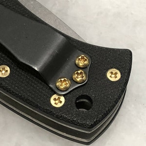 May include: Close-up of a black pocket knife with a clip and gold screws. The knife has a textured black handle and a silver blade.