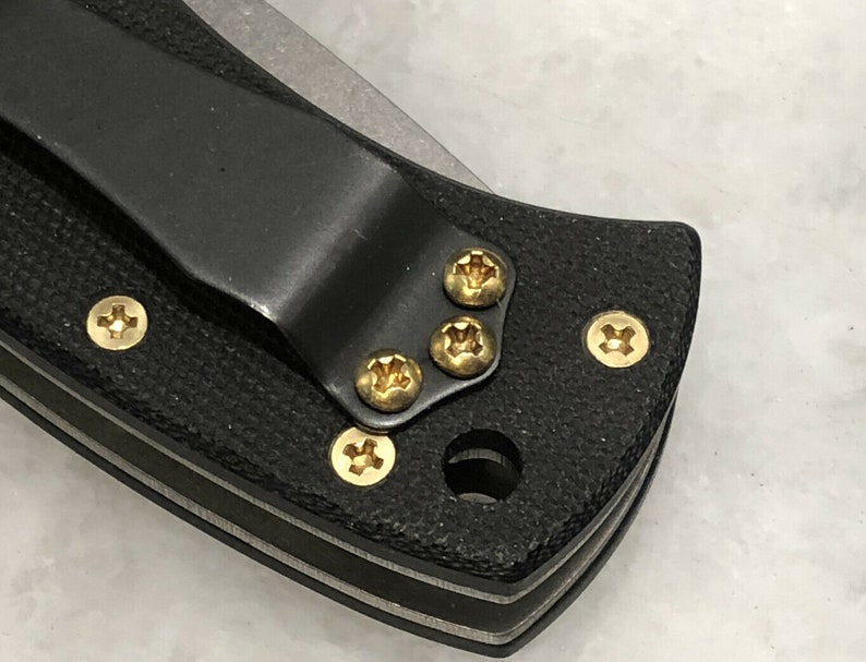 May include: Close-up of a black pocket knife with a clip and gold screws. The knife has a textured black handle and a silver blade.