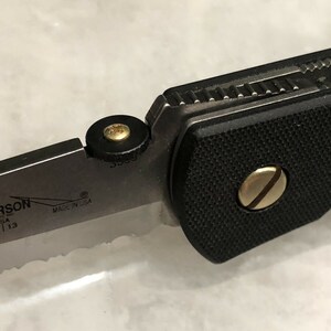 May include: Close-up of a black folding knife with a gold screw and a black and gold pivot. The knife has a partially serrated blade with the text "EMERSON USA 20 | 13 MADE IN USA" on the blade.