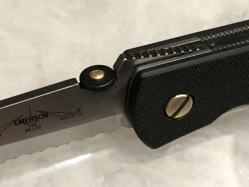 May include: Close-up of a black folding knife with a gold screw and a black and gold pivot. The knife has a partially serrated blade with the text "EMERSON USA 20 | 13 MADE IN USA" on the blade.