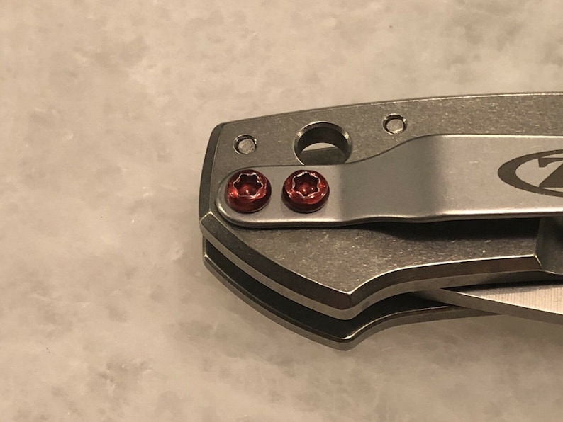 May include: Close-up of a silver pocket knife with a black handle and two red screws. The knife has a clip on the side and a logo on the handle.
