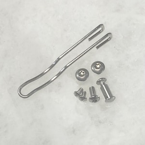 Könnte beinhalten: Eine Sammlung silberfarbener Hardware-Komponenten, darunter ein langer, gebogener Clip, zwei Rundkopfschrauben, mehrere kleine Schrauben und ein zylindrischer Pfosten. Die Artikel sind auf einer hellen Oberfläche angeordnet.