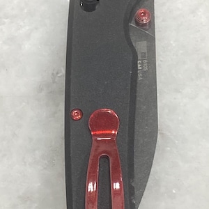 Pu&ograve; includere: Un coltello pieghevole nero con clip da tasca rossa e viti rosse. La lama &egrave; parzialmente visibile. Il coltello ha un manico strutturato e un piccolo bottone. Il coltello &egrave; contrassegnato "16105 CA USA".