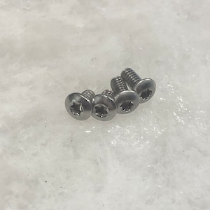 Puede incluir: Cuatro tornillos plateados con cabezas en forma de estrella están dispuestos en diagonal sobre una superficie blanca. Los tornillos tienen un vástago roscado y una cabeza plana con un patrón de estrella empotrado.