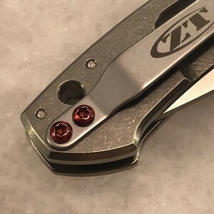 May include: Close-up of a silver pocket knife with a black clip and red screws. The knife has the text "ZT" on the clip.