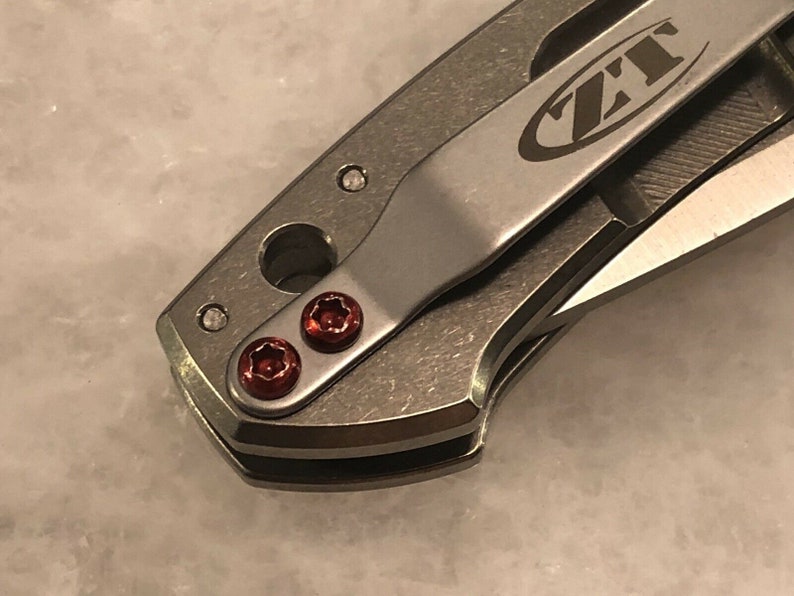 May include: Close-up of a silver pocket knife with a black clip and red screws. The knife has the text "ZT" on the clip.
