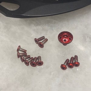 May include: A close-up of a set of small, red-colored screws with a star-shaped head. The screws are arranged in a semi-circle with a larger, red-colored screw with a star-shaped head in the center. The screws are all made of metal and have a shiny finish. The background is a white surface.