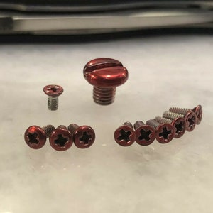 May include: A collection of red metal screws with a cross-shaped head. The screws are of various sizes, with the largest screw having a slightly rounded head.
