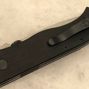 May include: Black folding knife with a black pocket clip and a textured black handle. The knife has a partial serrated blade.