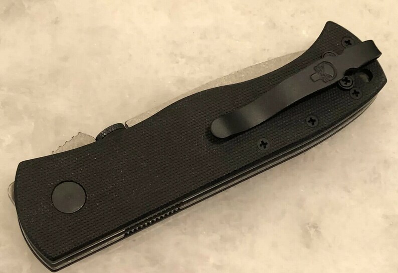 May include: Black folding knife with a black pocket clip and a textured black handle. The knife has a partial serrated blade.