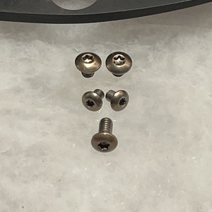 May include: Five silver-toned screws with a star-shaped head. The screws are arranged in a pattern on a white surface.