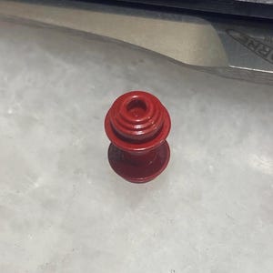 May include: A red metal bottle stopper with a circular design. The stopper has a small hole in the center.