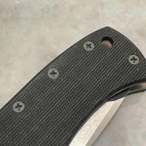 May include: Close-up of a black folding knife with a textured handle and four visible screws. The blade is partially open and the handle has a small hole.