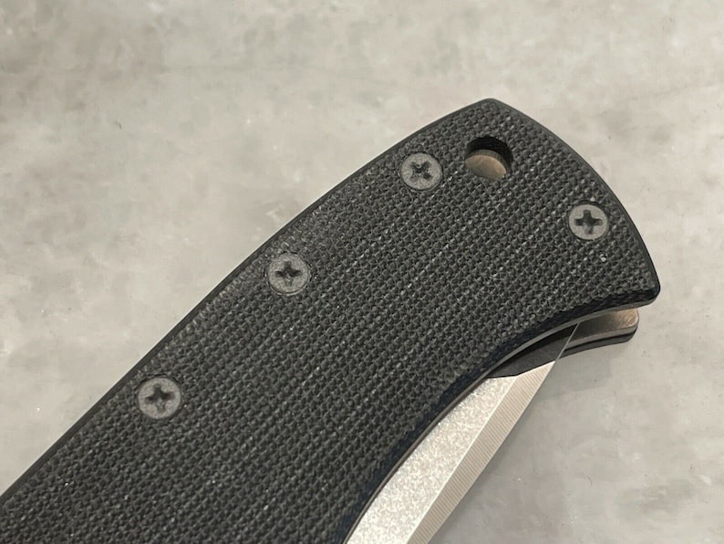 May include: Close-up of a black folding knife with a textured handle and four visible screws. The blade is partially open and the handle has a small hole.