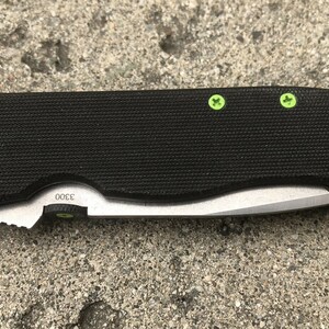 May include: Black folding knife with a green handle and green screws. The blade is partially open and the knife has a black textured handle. The knife has the number "3000" printed on the handle.
