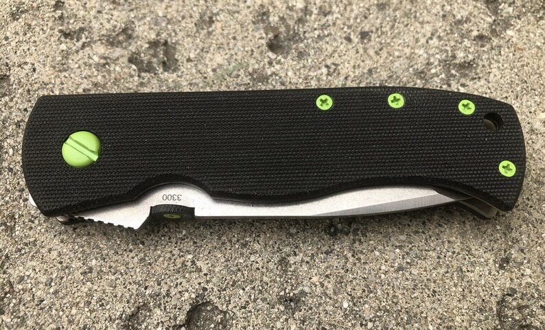 May include: Black folding knife with a green handle and green screws. The blade is partially open and the knife has a black textured handle. The knife has the number "3000" printed on the handle.