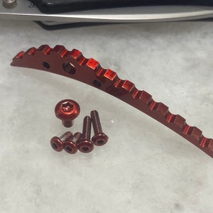 Puede incluir: Un conjunto de componentes de hardware metálicos rojos, que incluyen una pieza curva y estriada y varios tornillos. Los componentes están dispuestos sobre una superficie clara, con un cuchillo en el fondo. El hardware parece ser para un dispositivo mecánico.