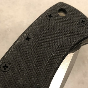 May include: Close-up of a black knife handle with a textured surface and four silver screws.