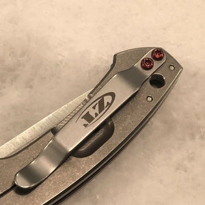 May include: A close-up of a silver pocket knife with a clip and red screws. The knife has the word "LZ" engraved on the clip.
