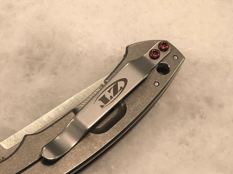 May include: A close-up of a silver pocket knife with a clip and red screws. The knife has the word "LZ" engraved on the clip.