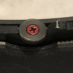 May include: Close-up of a black pocket knife with a red cross-shaped screw.