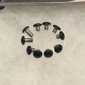 Peut inclure: Une collection de dix petites vis noires avec des filets argentés disposées en cercle. Les vis ont une tête plate et ronde avec une empreinte en forme d'étoile. Le fond est une surface claire et texturée.