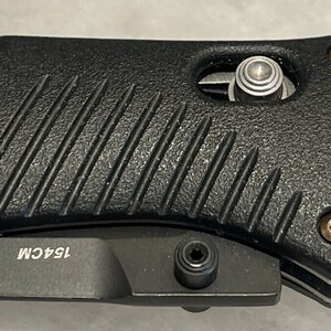 May include: Close-up of a black folding knife with a textured handle. The knife has a silver blade and a silver pocket clip. The handle has a silver screw and the text "154CM" is visible on the handle.