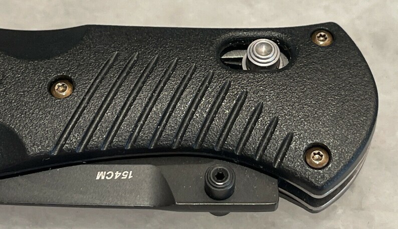 May include: Close-up of a black folding knife with a textured handle. The knife has a silver blade and a silver pocket clip. The handle has a silver screw and the text "154CM" is visible on the handle.