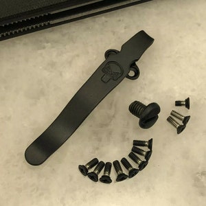 May include: Black metal pocket clip with a skull design and a set of black screws.
