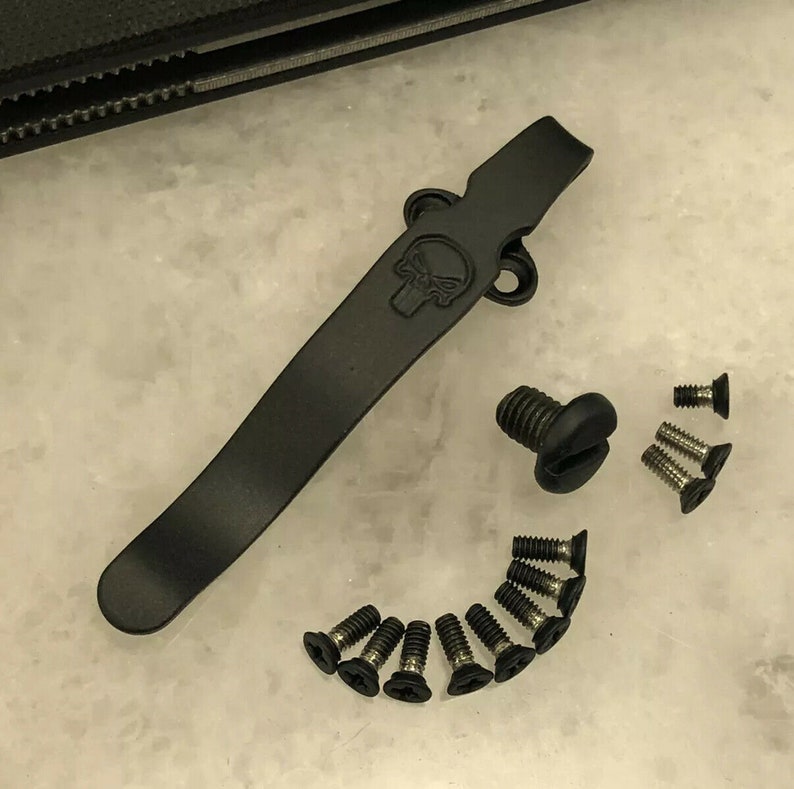 May include: Black metal pocket clip with a skull design and a set of black screws.