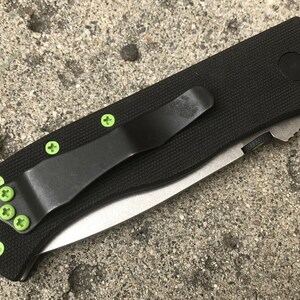 May include: A black folding knife with a green pocket clip and green screws. The knife has a black handle and a silver blade.