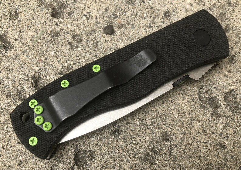 May include: A black folding knife with a green pocket clip and green screws. The knife has a black handle and a silver blade.