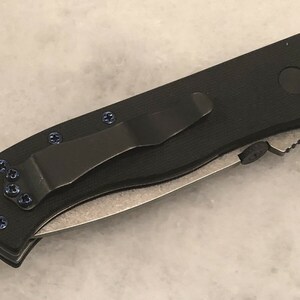 May include: Black pocket knife with a black clip and blue screws. The knife has a partially serrated blade.