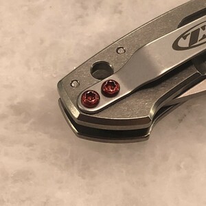 May include: Close-up of a silver pocket knife with a black ZT logo on the handle. The knife has two red screws on the handle.