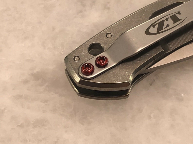 May include: Close-up of a silver pocket knife with a black ZT logo on the handle. The knife has two red screws on the handle.