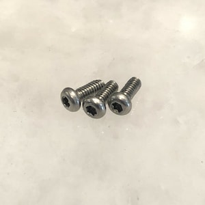 May include: Three silver-colored screws with a hex socket head. The screws have a threaded shaft and are arranged in a diagonal line. The heads are round and have a star-shaped indentation.