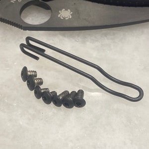 Può includere: Un coltello pieghevole nero con una clip da tasca e un set di viti nere. La lama del coltello è parzialmente visibile, con il logo Spyderco e il testo "D1N" sulla lama.