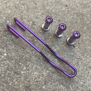 Op de afbeelding: Een metallic paarse U-vormige draadspeld en drie bijpassende metallic paarse cilindrische bevestigingsmiddelen met zeskantige schroefkoppen. De onderdelen hebben een gladde, glanzende afwerking op een grijs, gestructureerd oppervlak.