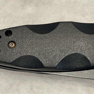 May include: Close-up of a black folding knife with a textured handle and two screws. The handle has a textured pattern and a small hole on the top.