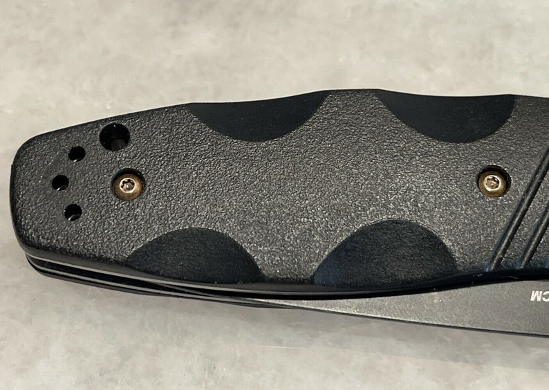 May include: Close-up of a black folding knife with a textured handle and two screws. The handle has a textured pattern and a small hole on the top.