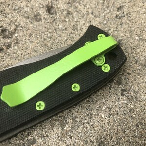 Pode incluir: Um canivete preto com um clip de bolso verde. O canivete tem um cabo texturizado e uma l&acirc;mina prateada.