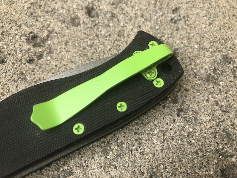 Pode incluir: Um canivete preto com um clip de bolso verde. O canivete tem um cabo texturizado e uma l&acirc;mina prateada.