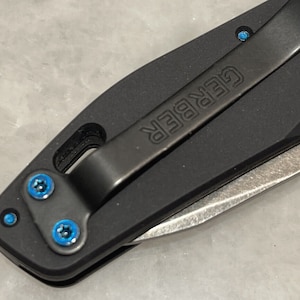Puede incluir: Primer plano de una navaja de bolsillo negra con una hoja plateada. La navaja tiene un clip de bolsillo con la palabra "GERBER" grabada en él. La navaja tiene cuatro tornillos azules.