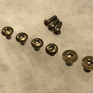 以下が含まれることがあります： 星型のくぼみがある、ブロンズ色のネジのコレクション。ネジは様々なサイズがあり、明るい大理石のような表面に配置されています。ネジの頭はわずかに丸みを帯びています。